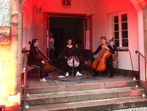 Zu sehen sind, im Halbkreis sitzend vor dem Eingang der Stadtbibliothek Meißen, drei Cello-Spielerinnen.